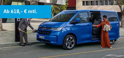 Blaue E-Caravelle Style in der Seitenansicht parkend am Stra&szlig;enrand beim Aufladen mit einer Frau und einem Mann daneben