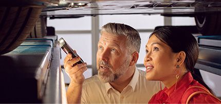 Eine Werkstattmitarbeiter zeigt einer Kundin mit einer Taschenlampe bei einem sich auf der Hebeb&uuml;hne befindenden Auto etwas am Boden des Autos